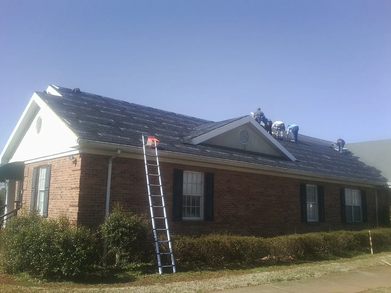 Crew tearing shingles off a brick ranch roof with a ladder set — Metal Roof Repair work by our crew in Walcott