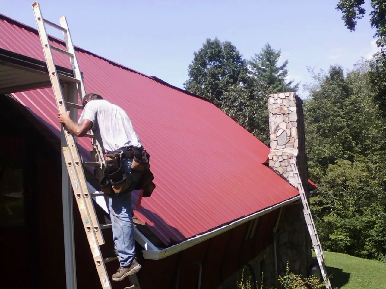 Roofer on a ladder beside a red metal roof and stone chimney — Soffit & Fascia Repair by Roofing Brothers in Walcott